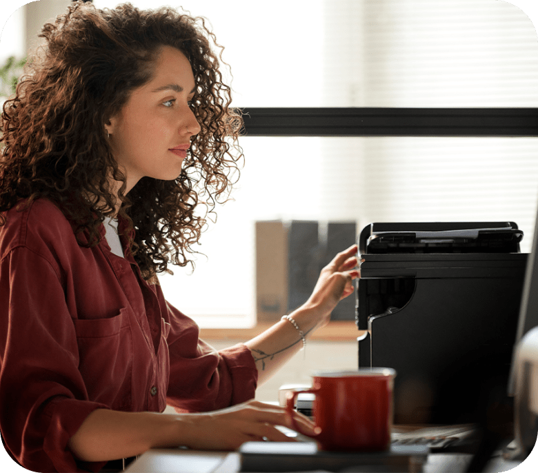 Mujer sentada en un escritorio utilizando la impresora con el pelo castaño rizado y camisa roja